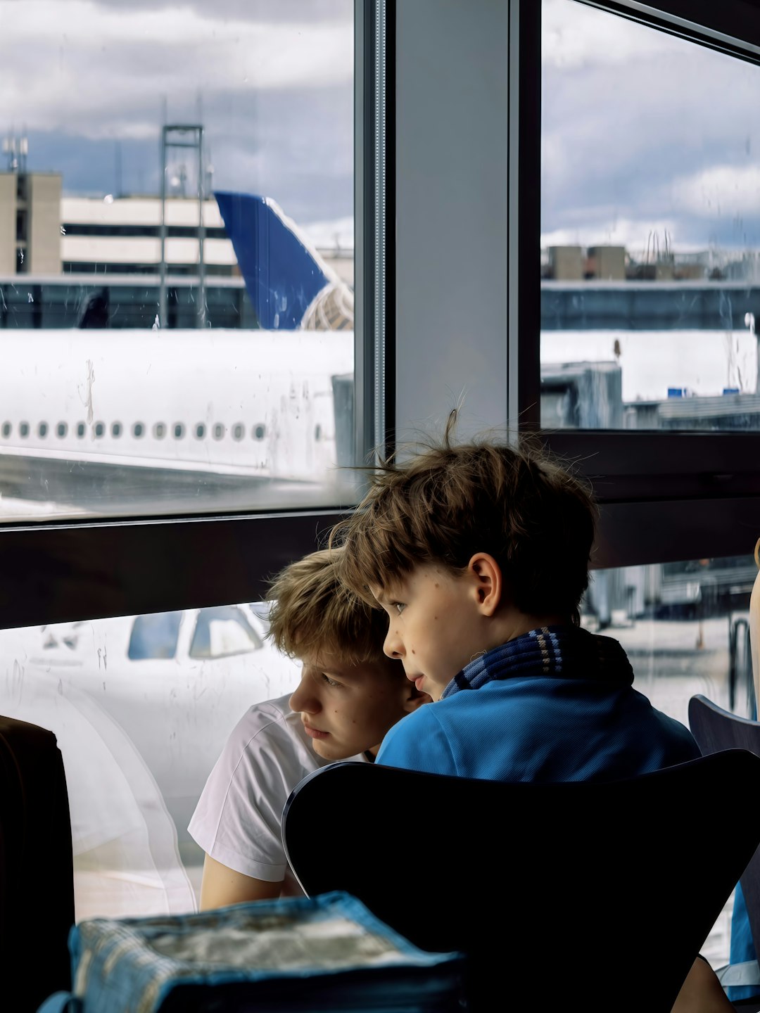 Two boys look out airport window at airplane.